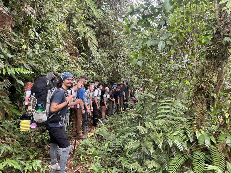 A group of people is hiking on a trail through a lush, green forest. The trail is narrow, and the hikers are walking in a line. The person in the front is wearing a large backpack. The forest is dense with vegetation.